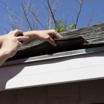 Hand lifting up roofing shingles to inspect storm damage.